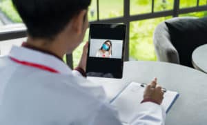 The doctor's back view is communicating with the patient wear a mask via tablets. Doctors are using Telemedicine technology to interact with patients via video conference for keep social distancing